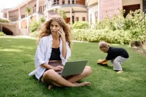 Woman working on a laptop while talking on the phone, with a child playing nearby in a garden setting