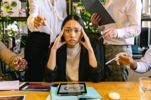 Woman sitting at a desk with a stressed expression, surrounded by people pointing and holding various objects, associated with 'When a Texas Airbnb Host’s Chores List Goes Viral' article – by Open Air Homes.