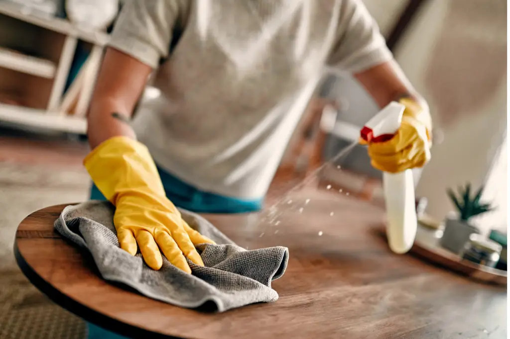 Close-up of a person wearing yellow gloves cleaning a wooden surface with a cloth and spray bottle, emphasizing cleanliness - by Open Air Homes.