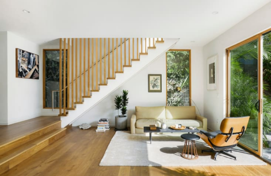 Living area with wooden flooring, staircase with wooden railings, sofa, and large windows - by Open Air Homes.