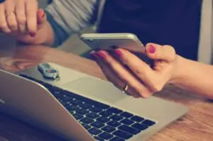 Person holding a smartphone with both hands, with a laptop and keychain in the background, representing the use of technology in managing Airbnb rentals - by Open Air Homes.