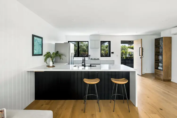 Modern kitchen with white island, bar stools, and gas cooktop at Westminster Bungalow - by Open Air Homes.Kitchen with white cabinets and black countertops at Westminster Bungalow - by Open Air Homes.