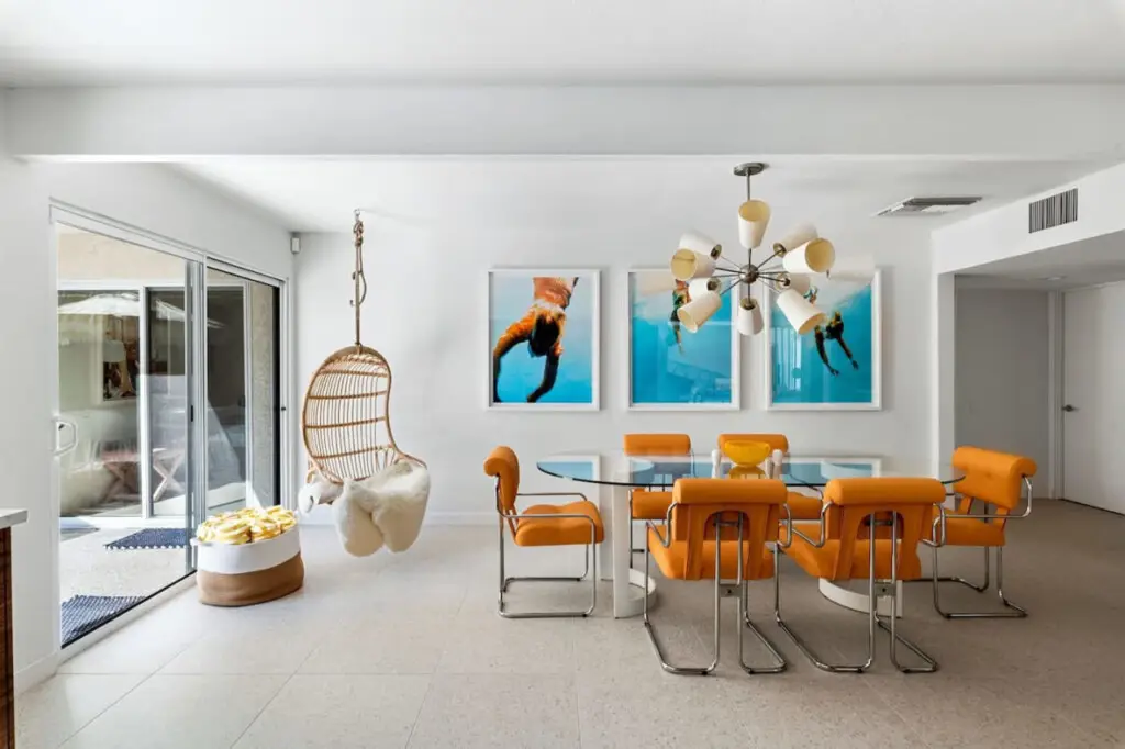 Dining area with glass table, orange chairs, hanging wicker chair, and artwork at Monterey Manor in Palm Springs - Designer Home by Open Air Homes