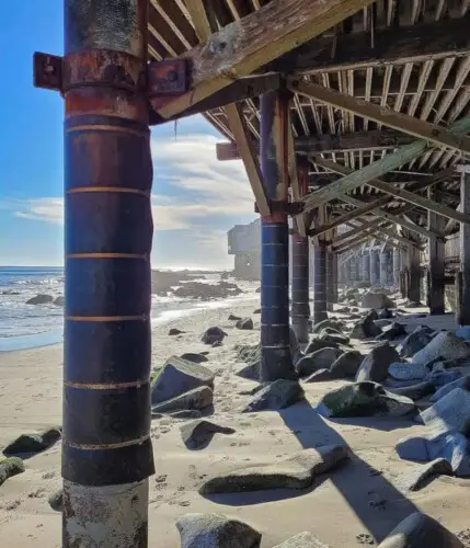 View under pier with sand, rocks, and ocean at Breathe Malibu - Ocean Front Condo w/ Beach Access by Open Air Homes.