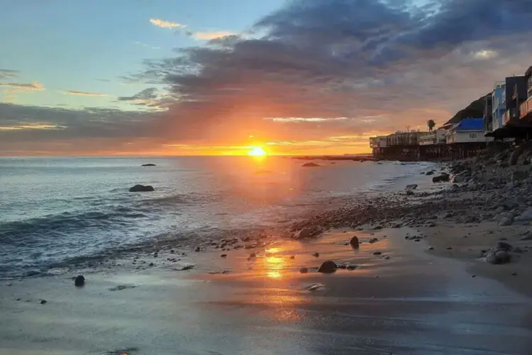 Sunset over the ocean with houses along the shoreline at Breathe Malibu - Ocean Front Condo w/ Beach Access by Open Air Homes.