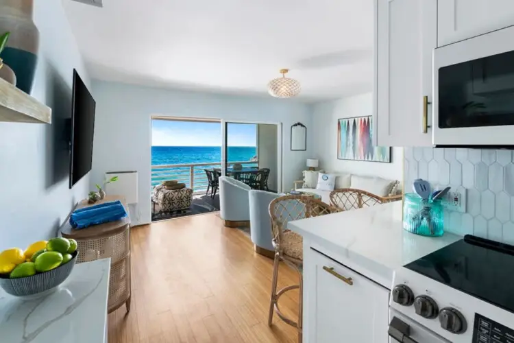 Interior view of living room and kitchen with open door leading to balcony overlooking the ocean at Breathe Malibu - Ocean Front Condo w/ Beach Access by Open Air Homes.