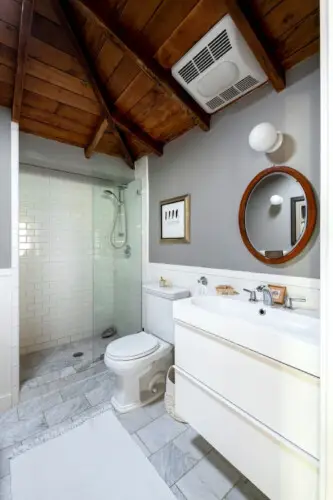 Bathroom with shower area, toilet, sink, round mirror, and wooden ceiling at Hillside Hideaway in Topanga Canyon by Open Air Homes.