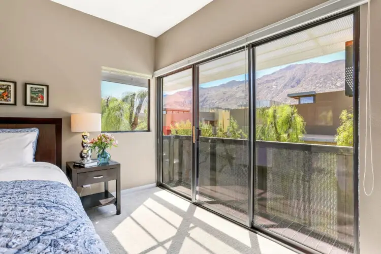 Bedroom with bed, side table, lamp, and sliding glass doors leading to a balcony with mountain view at La Mirage gated community in Palm Springs – by Open Air Homes.