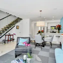 Open living space with modern staircase, seating area with blue sofa and chairs, and dining area adjacent to kitchen with white cabinetry and blue accent wall at La Mirage - by Open Air Homes