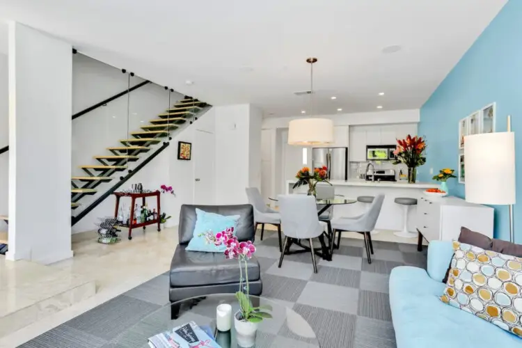 Open living space with modern staircase, seating area with blue sofa and chairs, and dining area adjacent to kitchen with white cabinetry and blue accent wall at La Mirage - by Open Air Homes