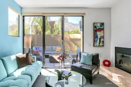 Living room with blue sofa, glass coffee table, fireplace, and sliding glass door to outdoor seating area at La Mirage - by Open Air Homes