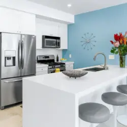 Modern kitchen with white cabinetry, stainless steel appliances, blue accent wall, and white countertop with bar stools at La Mirage - by Open Air Homes