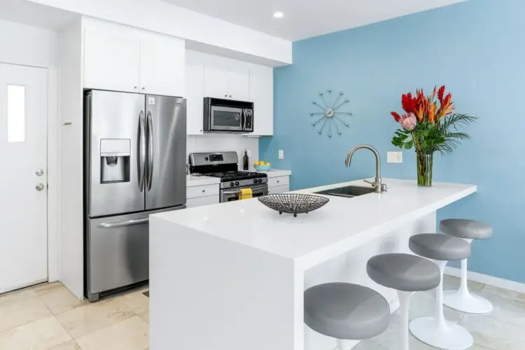 Modern kitchen with white cabinetry, stainless steel appliances, blue accent wall, and white countertop with bar stools at La Mirage - by Open Air Homes