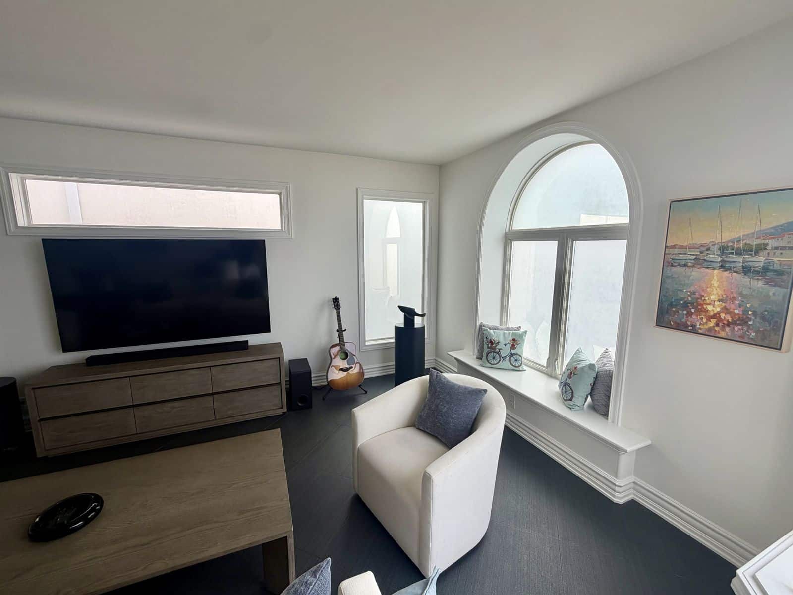 Living area with a 75 inch smart TV, swivel chair, arched window seat and harbor painting