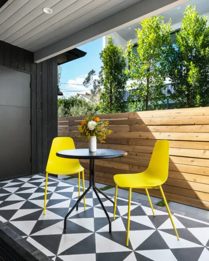 Outdoor patio with small table and yellow chairs, adjacent to wooden fence and greenery – by Open Air Homes.