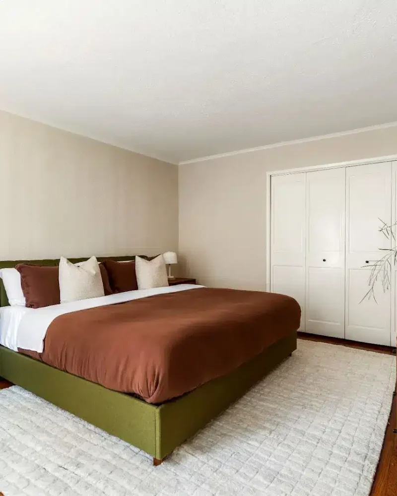 Bedroom with bed, brown bedding, wooden dresser, and large plant creating a cozy and inviting atmosphere – by Open Air Homes.